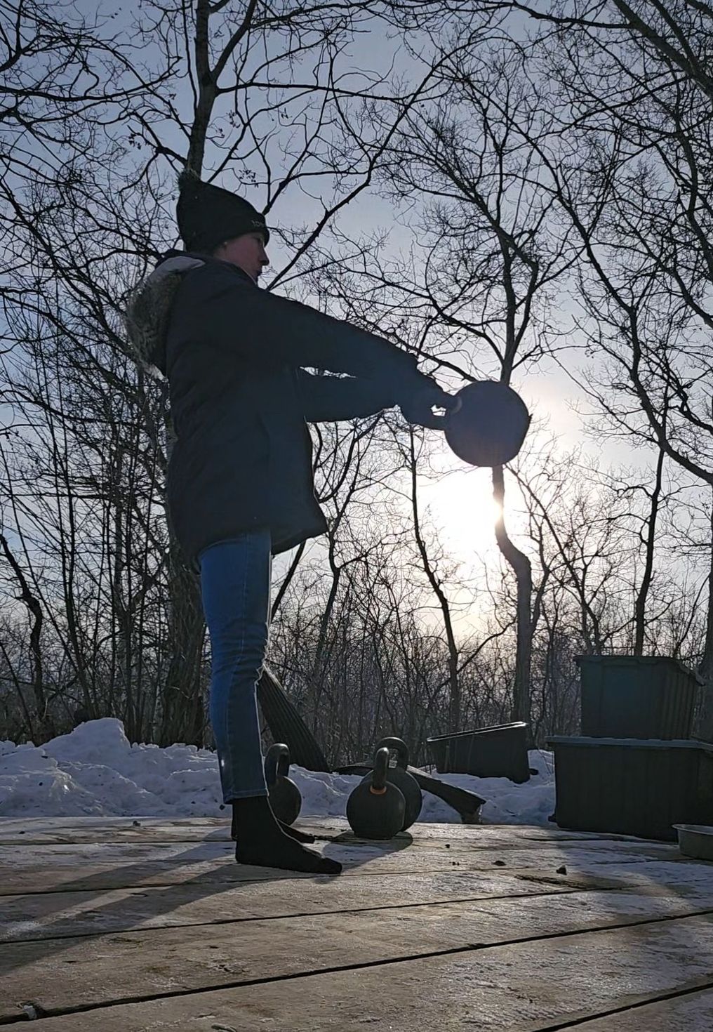 A person in winter clothes performs a kettlebell swing on a snow-covered deck against a backdrop of bare trees and sunlight.