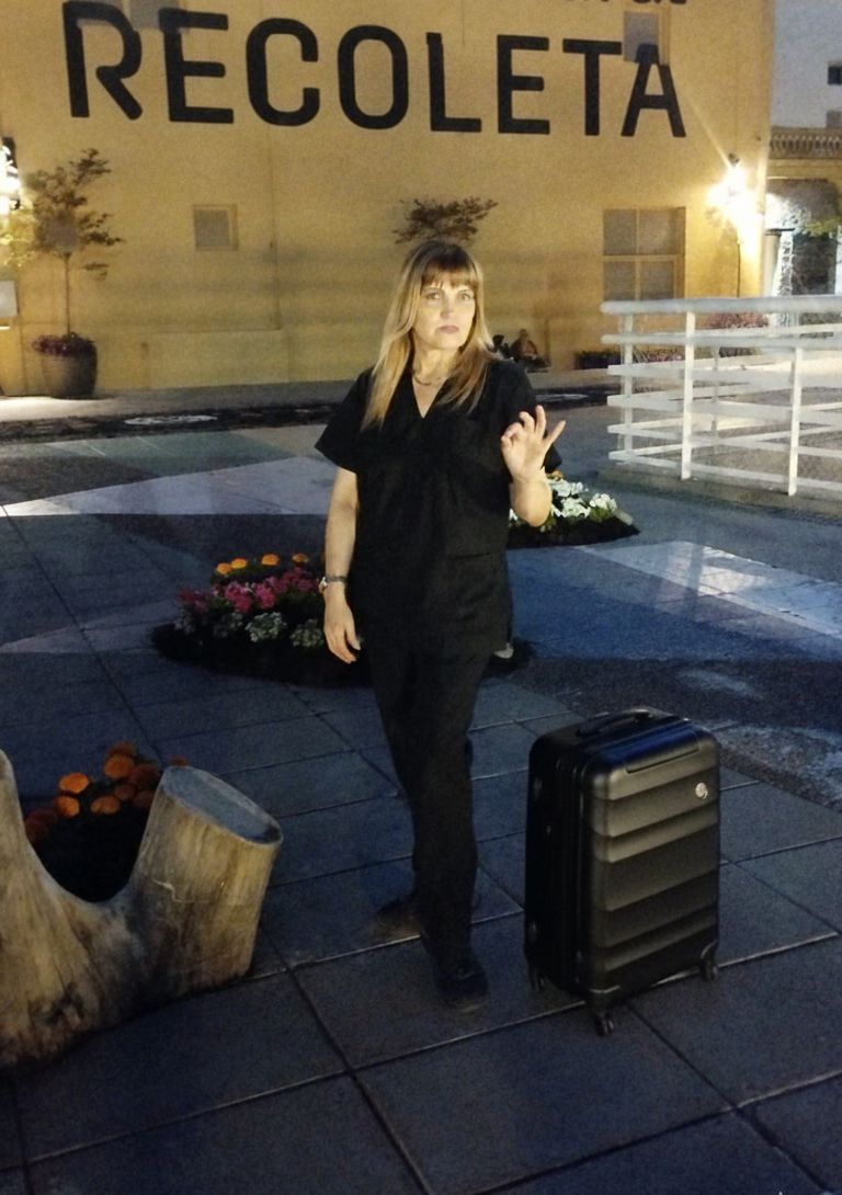 Una mujer vestida con bata negra gesticula cerca de una maleta; en la pared del fondo se lee 