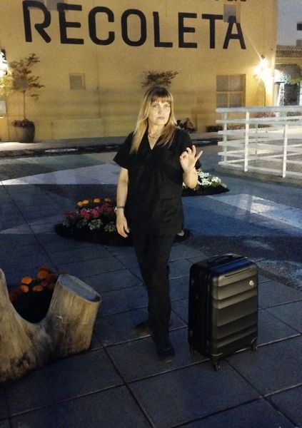 Una mujer vestida con bata negra gesticula cerca de una maleta; en la pared del fondo se lee 