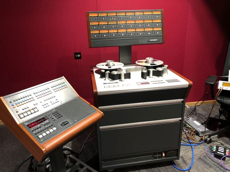 Vintage multi-track tape recorder in a studio setting. Grey and brown control panel, red wall.