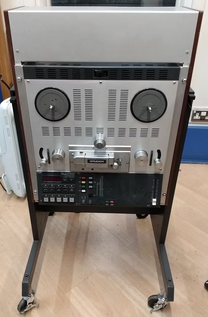 Large reel-to-reel tape recorder on a wheeled stand, wooden cabinet, silver faceplate, black controls.