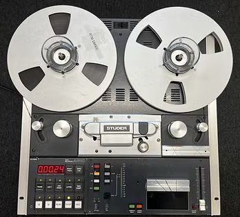 Vintage Studer reel-to-reel tape recorder, with two silver reels. Black console with buttons and a VU meter.