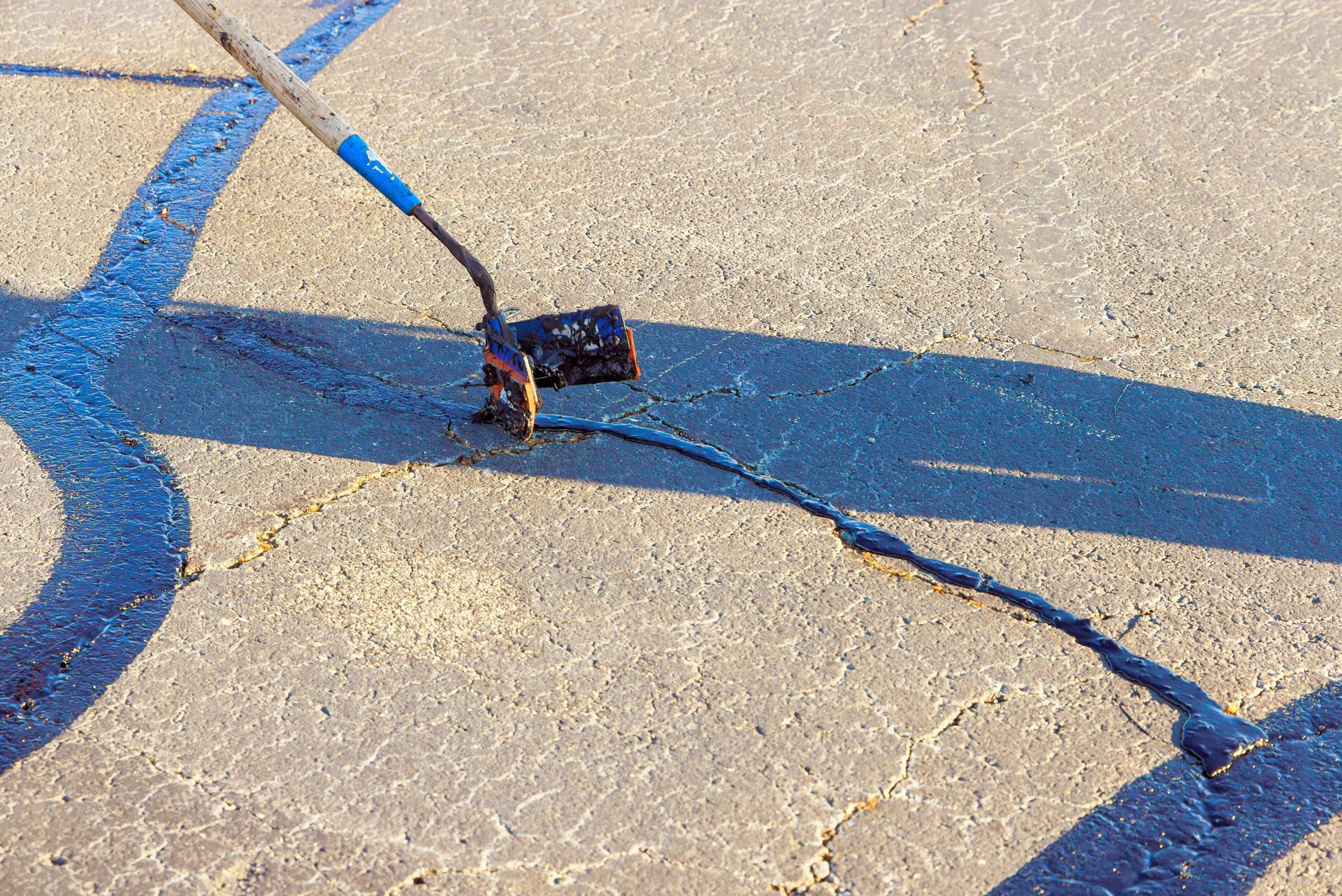 Applying sealant to a crack in asphalt with a long-handled tool; sunny day.
