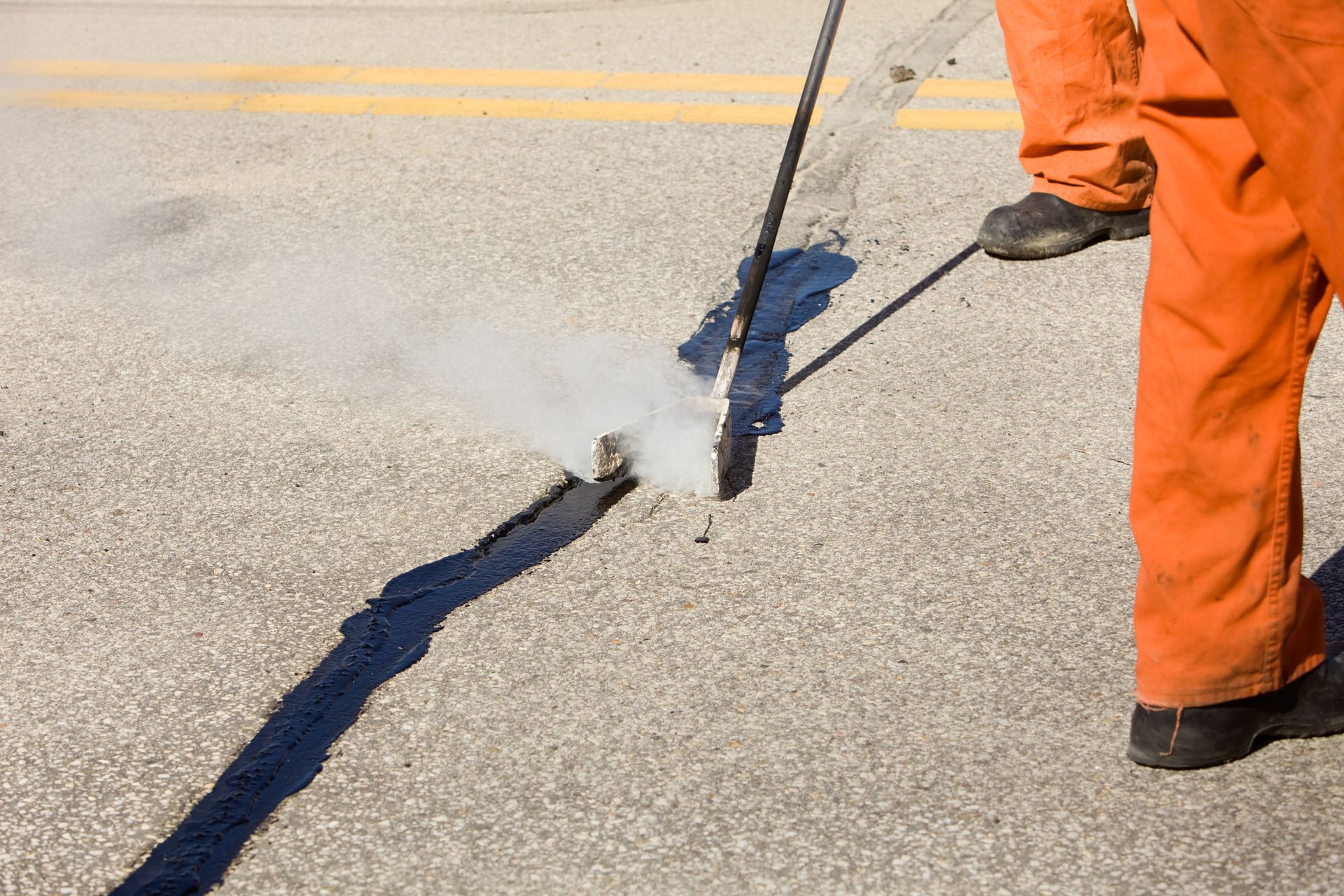 Person in orange jumpsuit seals crack in asphalt road with smoke emitting from the repair.