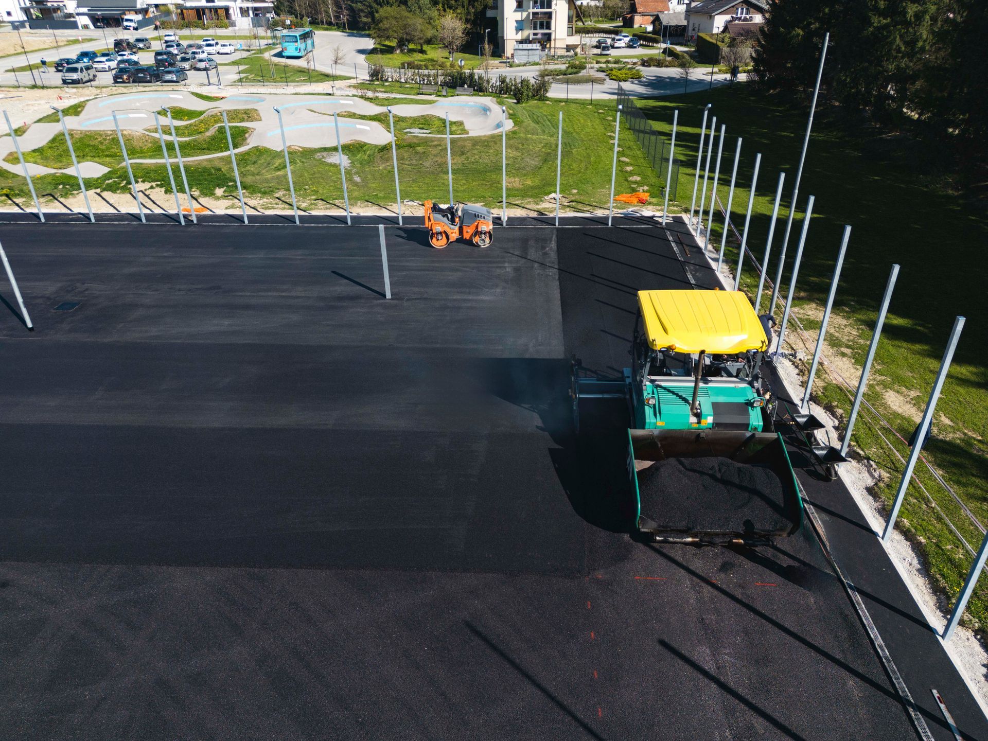 Asphalt paving in progress; a yellow and green machine smoothing dark pavement, surrounded by metal poles.