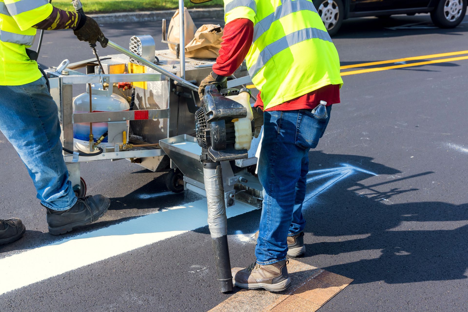 Two road workers painting a white line on asphalt, wearing safety vests.