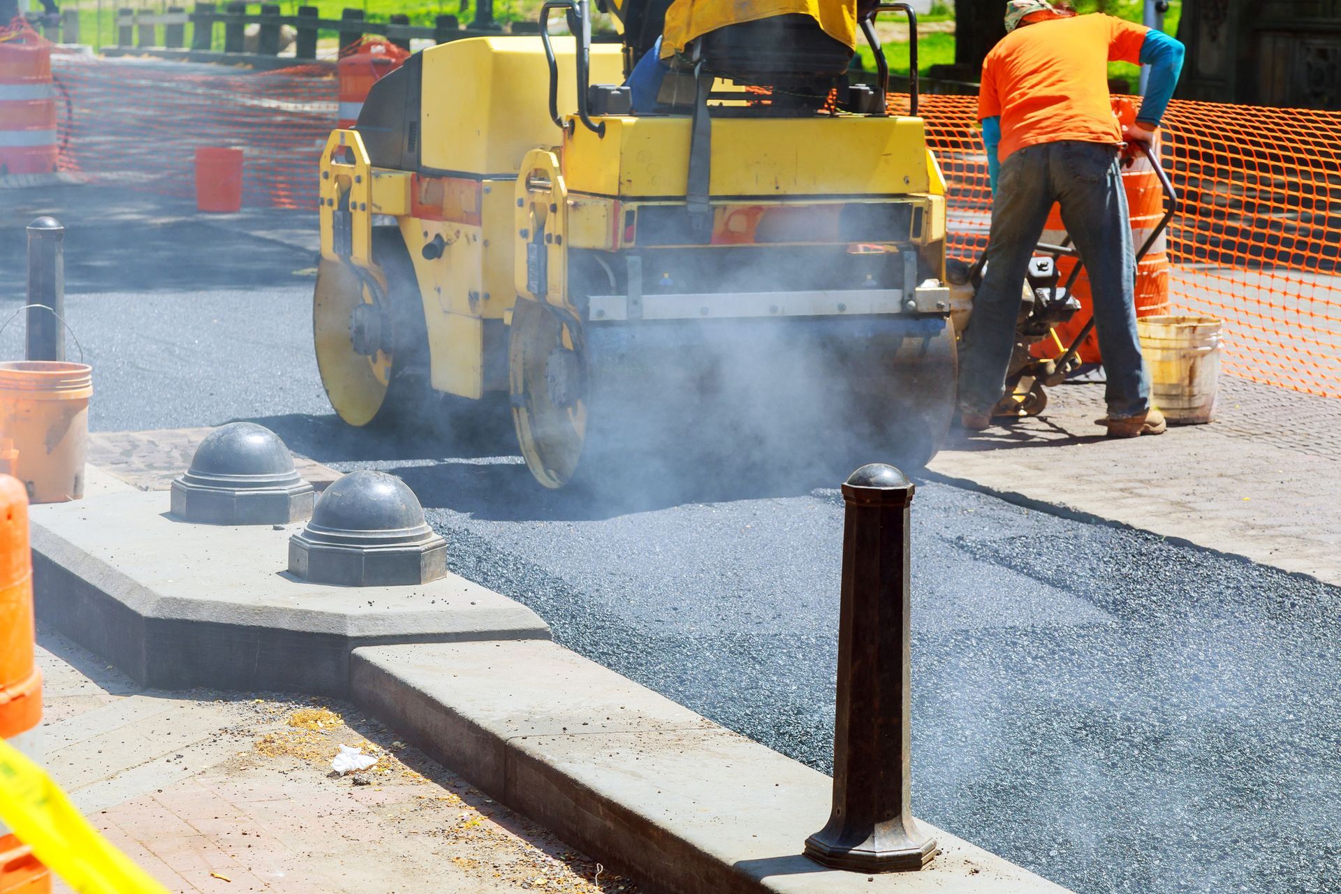Road paving in progress; yellow steamroller compacts asphalt; worker in orange shirt uses a vibratory plate compactor.