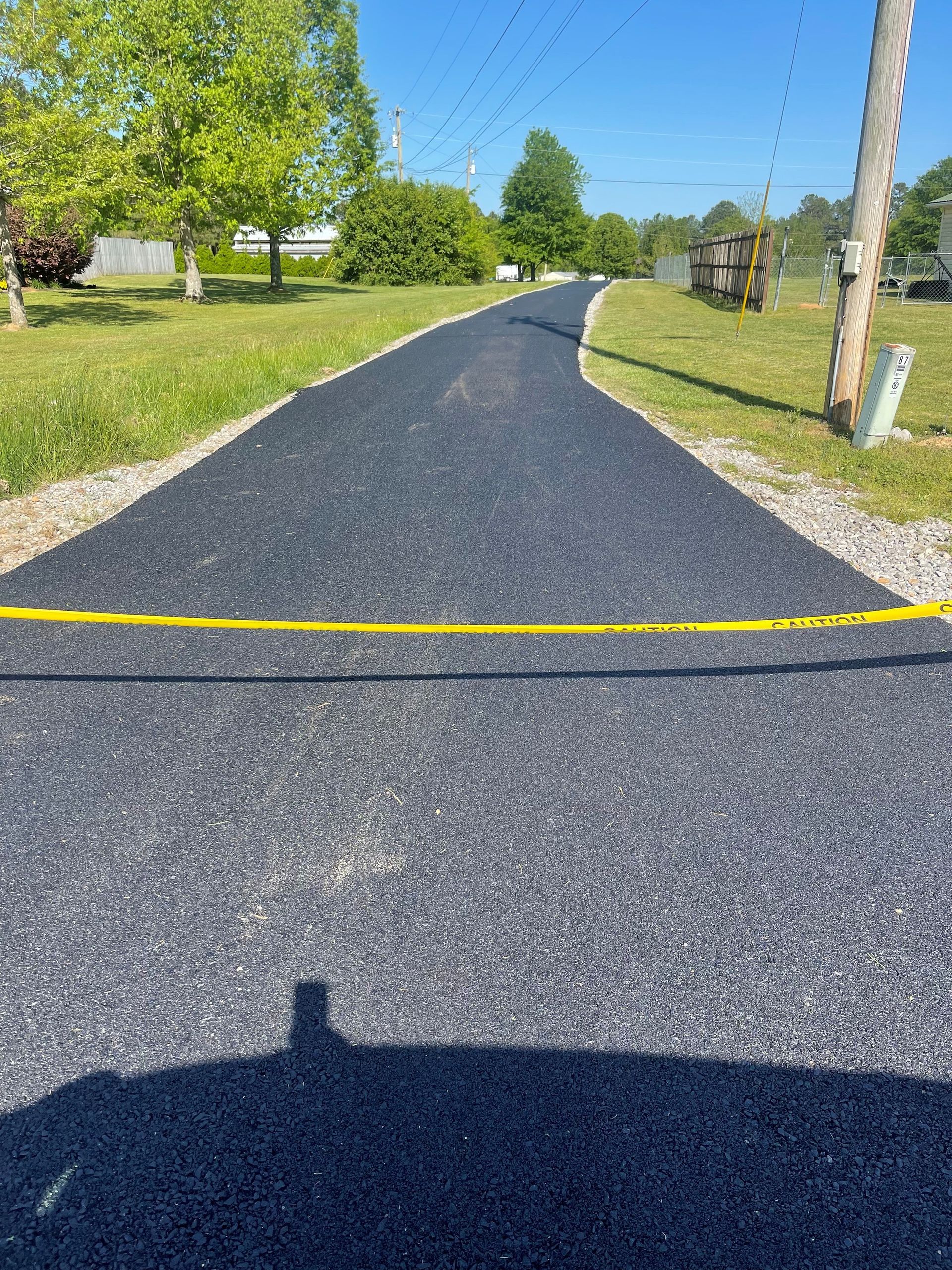 Newly paved asphalt driveway with yellow caution tape, leading towards trees and houses on a sunny day.