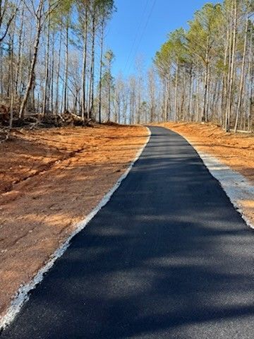 Paved pathway through a wooded area with trees on either side and reddish-brown dirt.