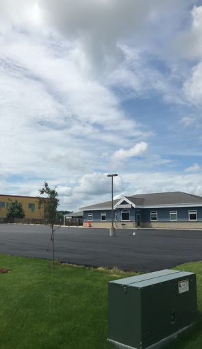 Blue building with white trim under a cloudy blue sky. Black asphalt parking lot and green grass in the foreground.