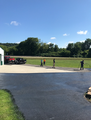 Workers sealcoating a paved driveway in front of a building, green field and trees under a blue sky.