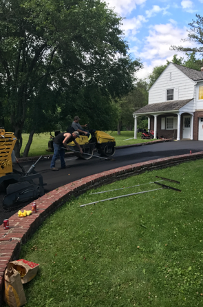 Workers paving a driveway with a roller, beside a brick-lined flower bed and a two-story house.