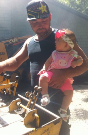 Man in a cap and black tank top holds a baby in pink clothes near construction equipment.