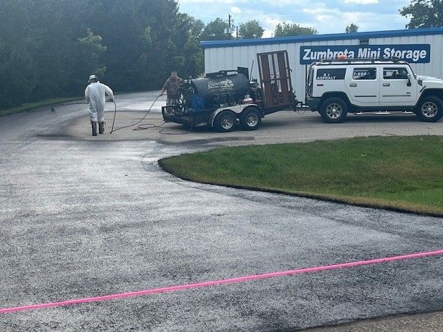 Person using a roller to apply dark coating to a concrete surface; they wear gloves, jeans, and a blue jacket.