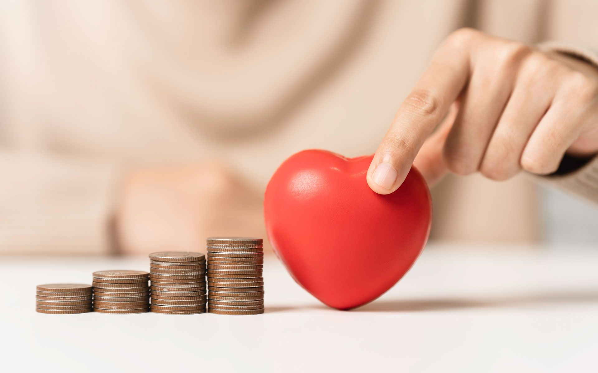 Person's hand touching a red heart, with stacks of coins in front, on a white surface.
