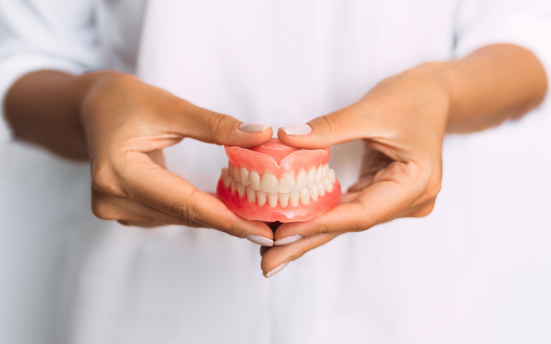 Hands holding a full set of dentures, pink base with white teeth.