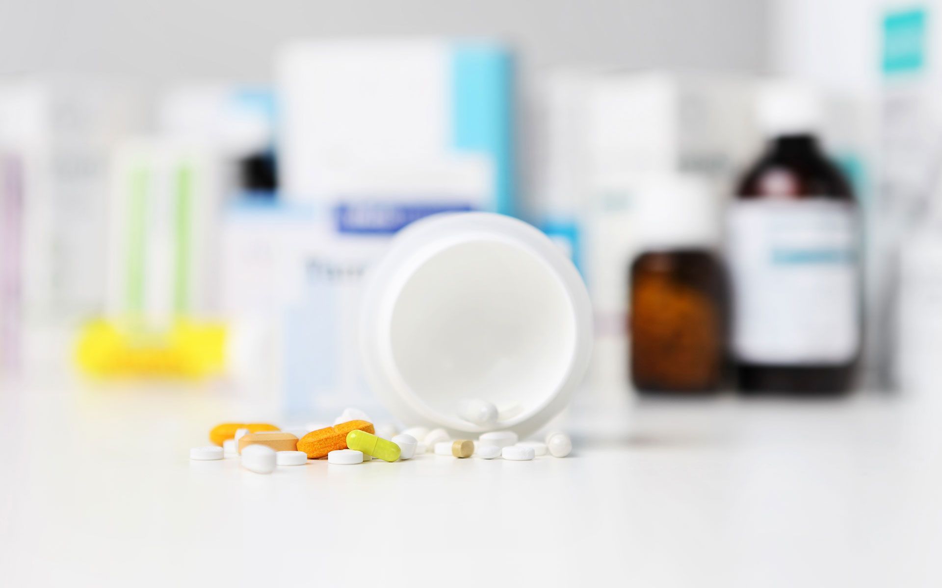 Open white pill bottle with assorted pills spilled on white surface.