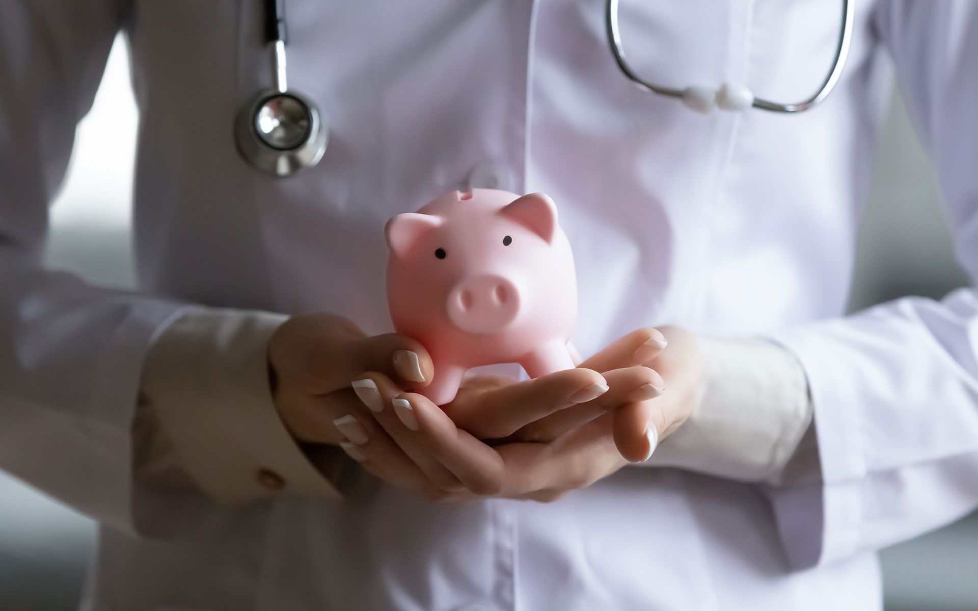 Doctor holding pink piggy bank, stethoscope around neck, white coat.