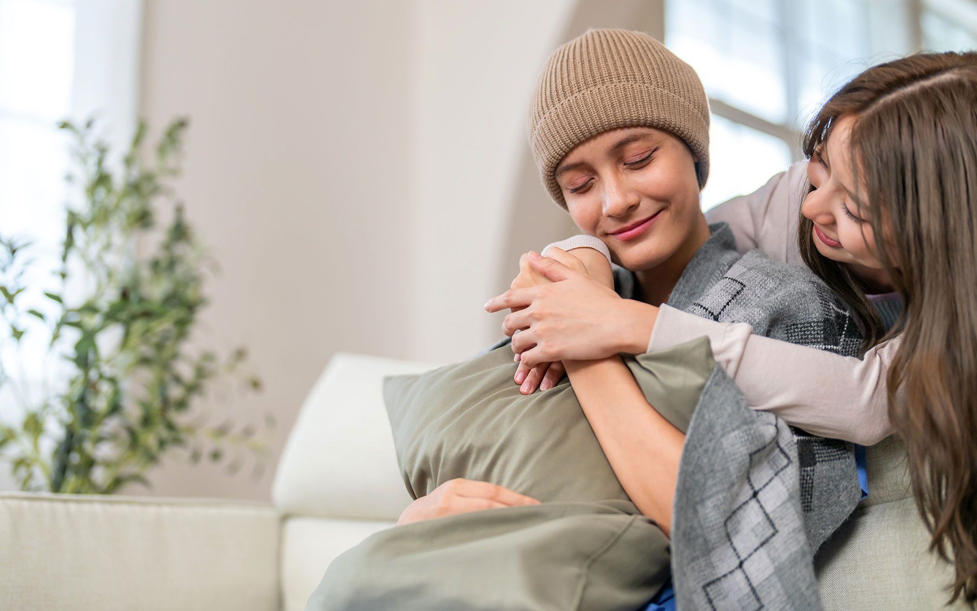 Woman with cancer in beanie embraces her daughters on a couch, showing support.