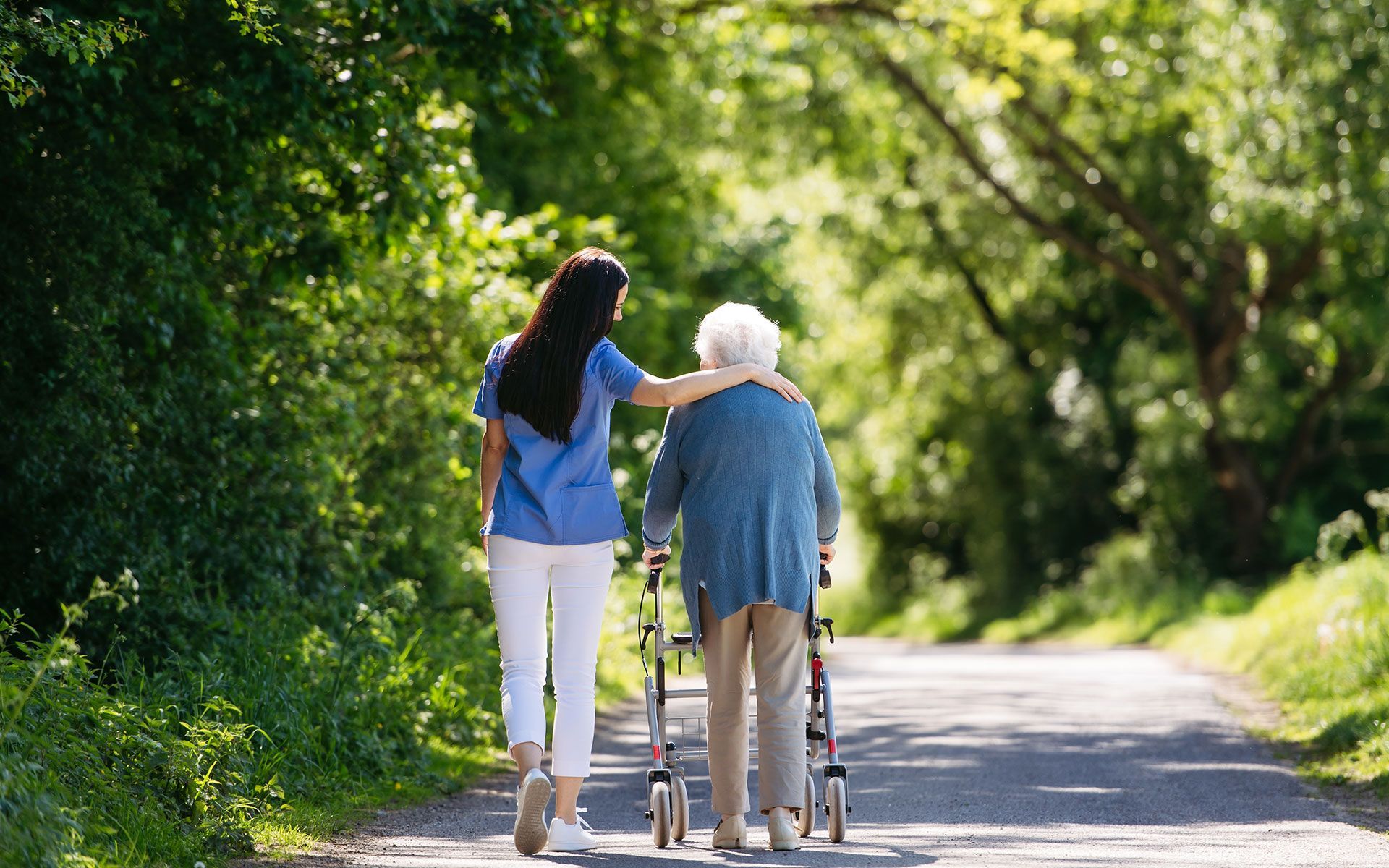 Woman in scrubs assists elderly woman with walker down a path lined with trees.