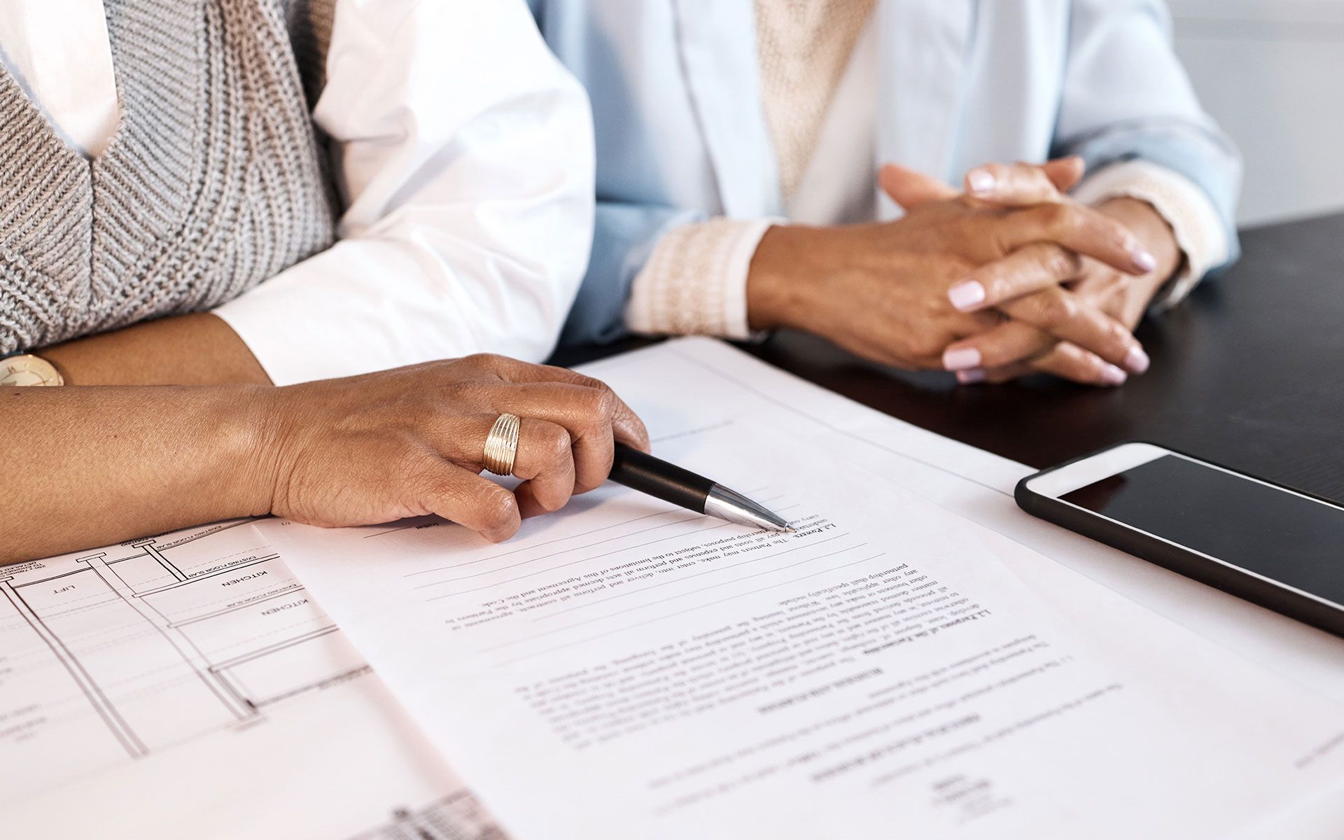 Two people reviewing documents, one holding a pen. Papers and a phone are on the table.