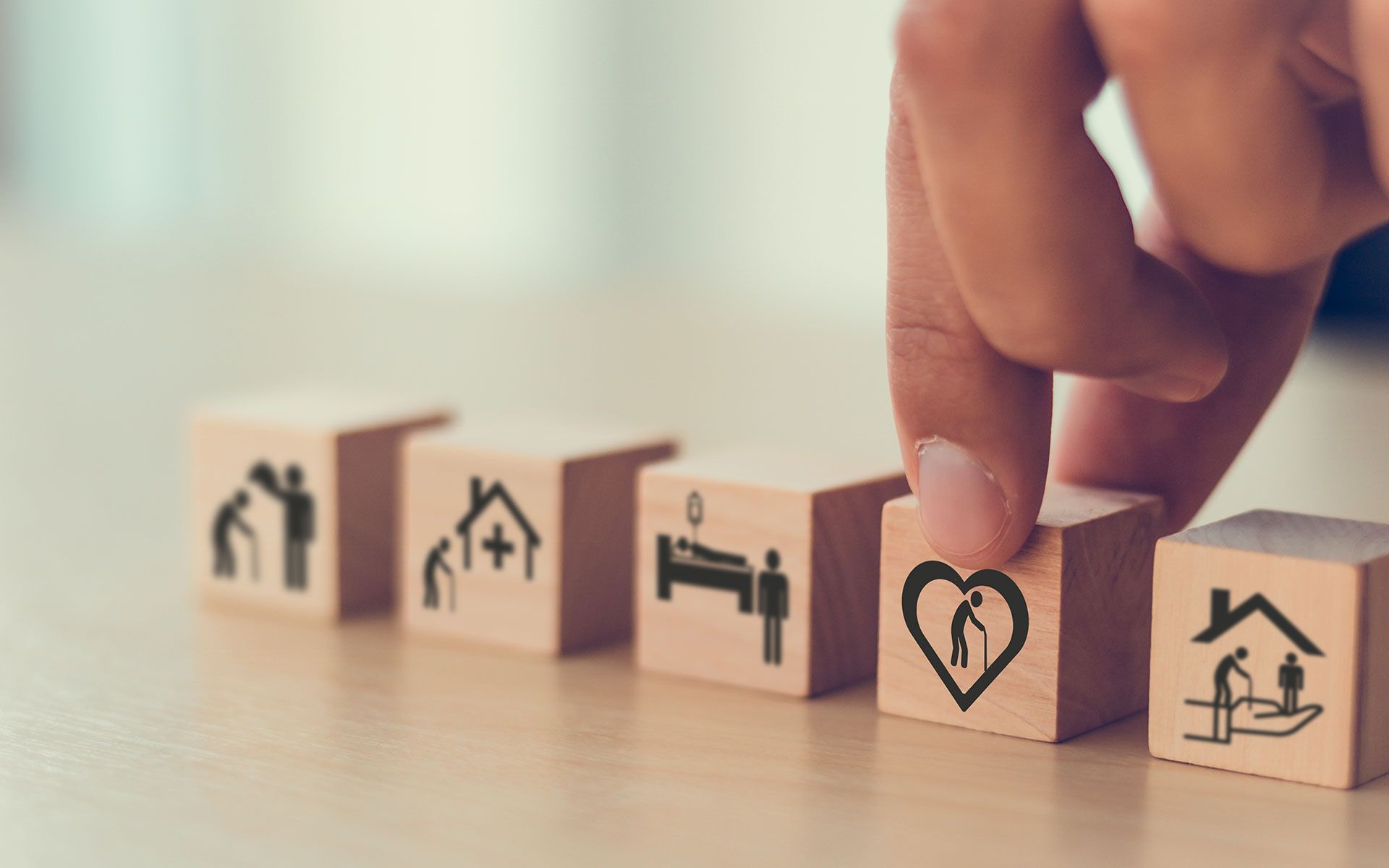 Hand arranging wooden blocks with care symbols: elderly care, home, hospital bed, heart, and home with elderly person.