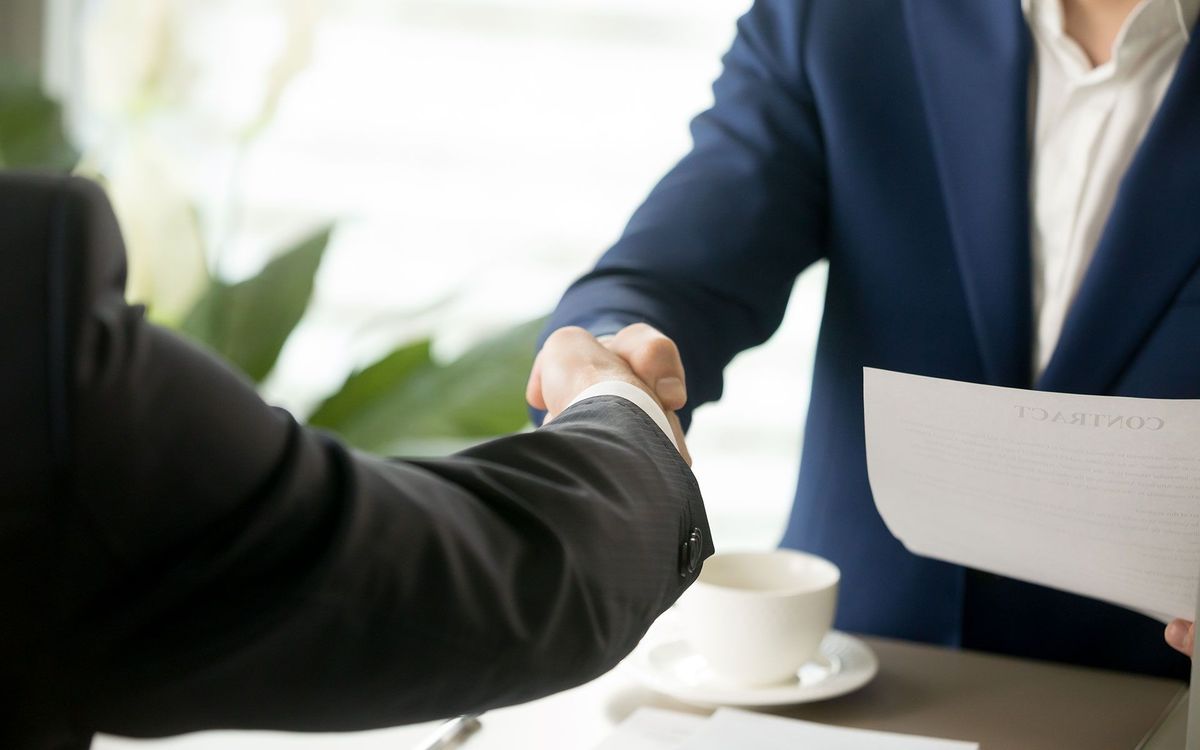 Two men in suits shaking hands, signifying a deal. One holds paperwork near a cup.