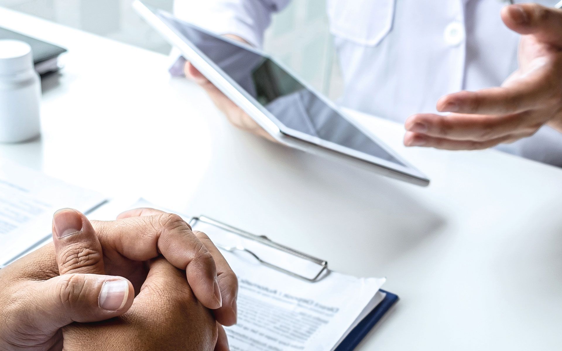 Doctor in white coat showing information on a tablet to a patient, hands clasped, at a desk.