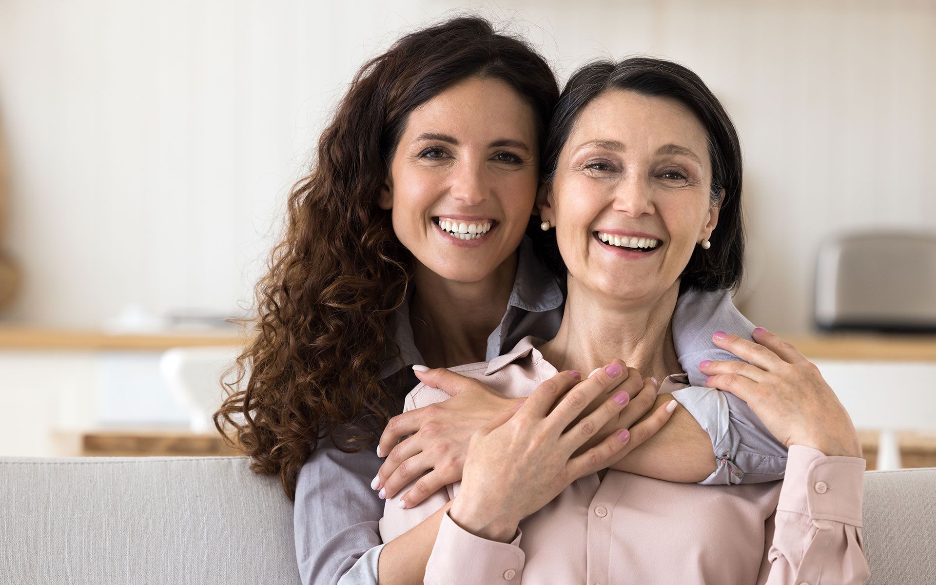 Woman embraces older woman; both smile, seated indoors.