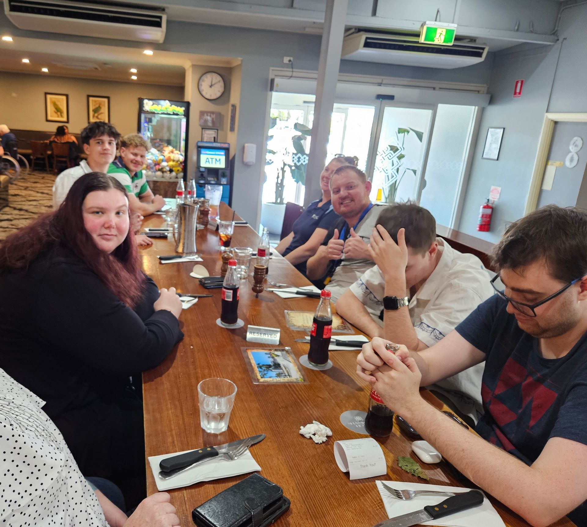 A Group Of People Sit At A Long Wooden Dining Table In A Restaurant, Eating And Socializing — Advanced Therapy Mackay In Mackay, QLD 