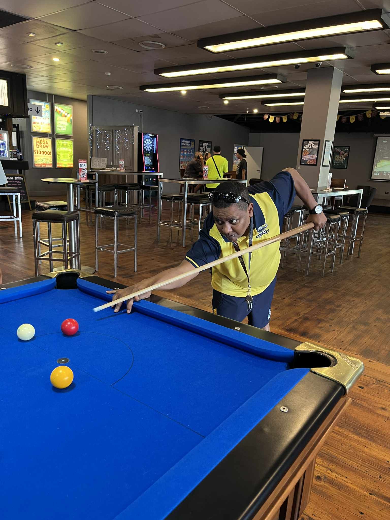 A person in a yellow vest leans over a pool table in a lounge, aiming a cue at a white, red, and yellow ball — Advanced Therapy Mackay In Mackay, QLD 