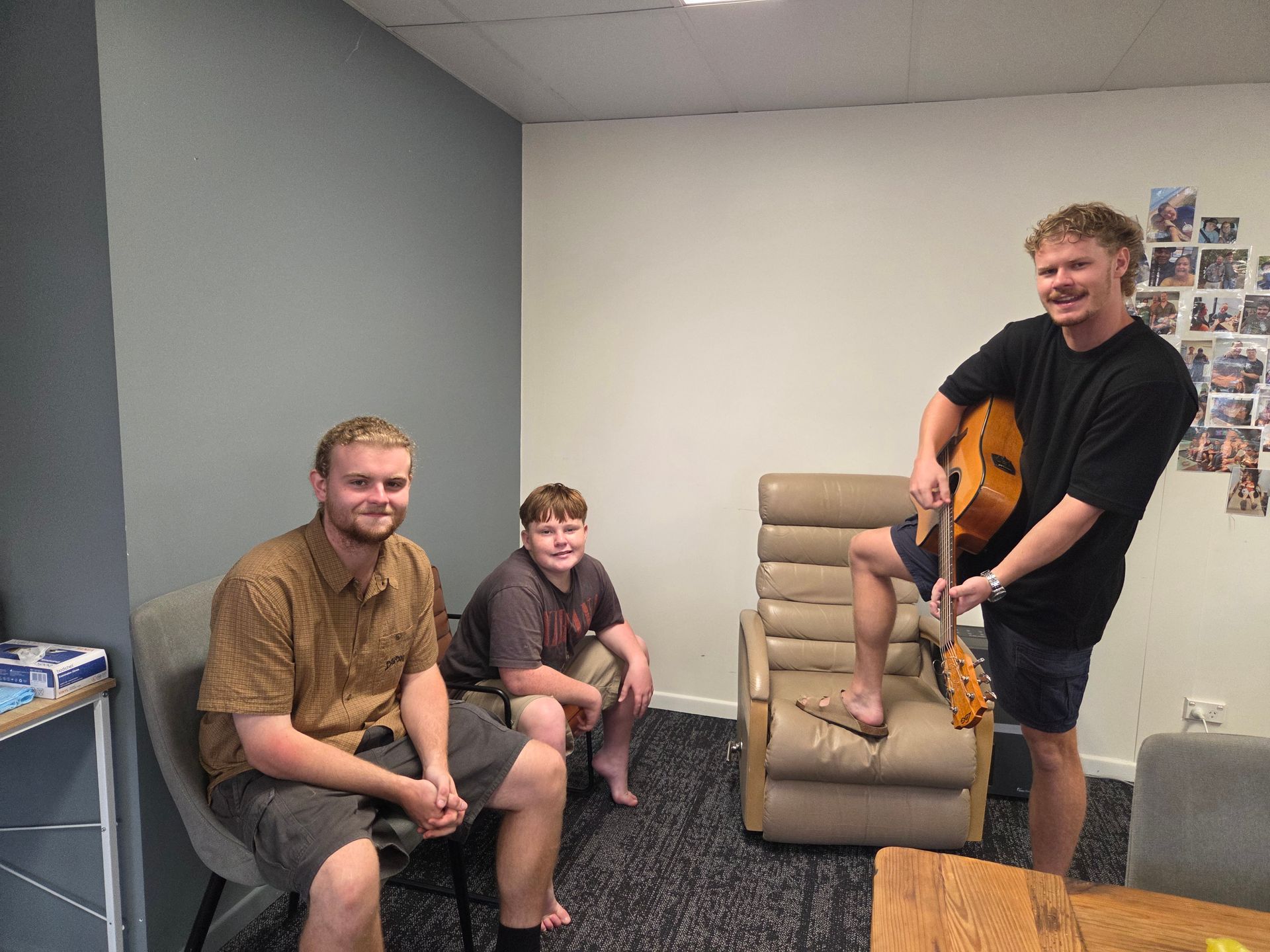 Three people are in a room: one sitting in a gray chair, one on a stool, and one standing while holding an acoustic guitar — Advanced Therapy Mackay In Mackay, QLD 