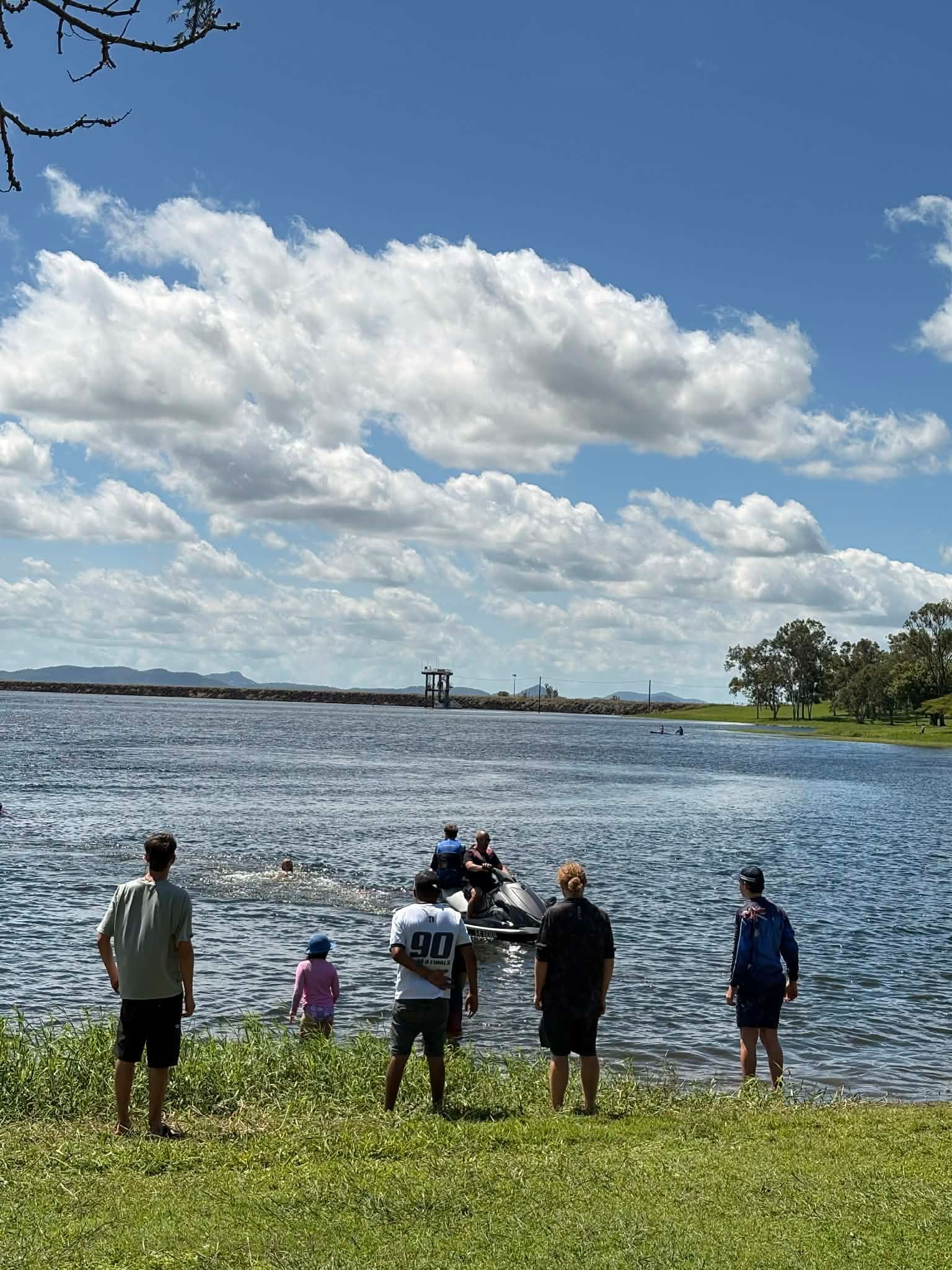 A group of people standing on a grassy shore looking out at a small boat on a blue lake under a partly cloudy sky — Advanced Therapy Mackay In Mackay, QLD 