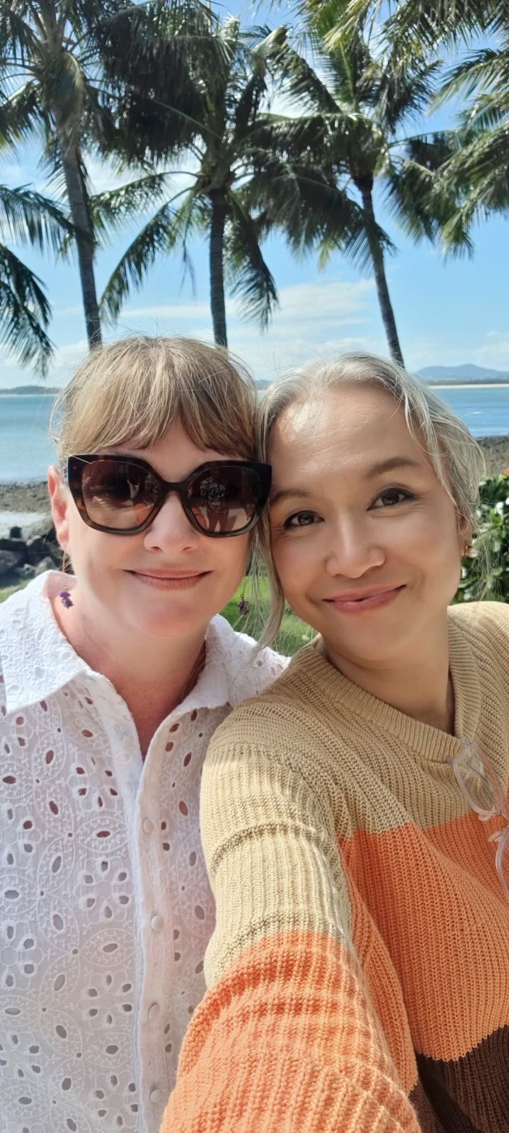 Two Smiling People Pose For A Selfie Outdoors With Palm Trees, A Calm Ocean, And A Clear Blue Sky In The Background — Advanced Therapy Mackay In Mackay, QLD 