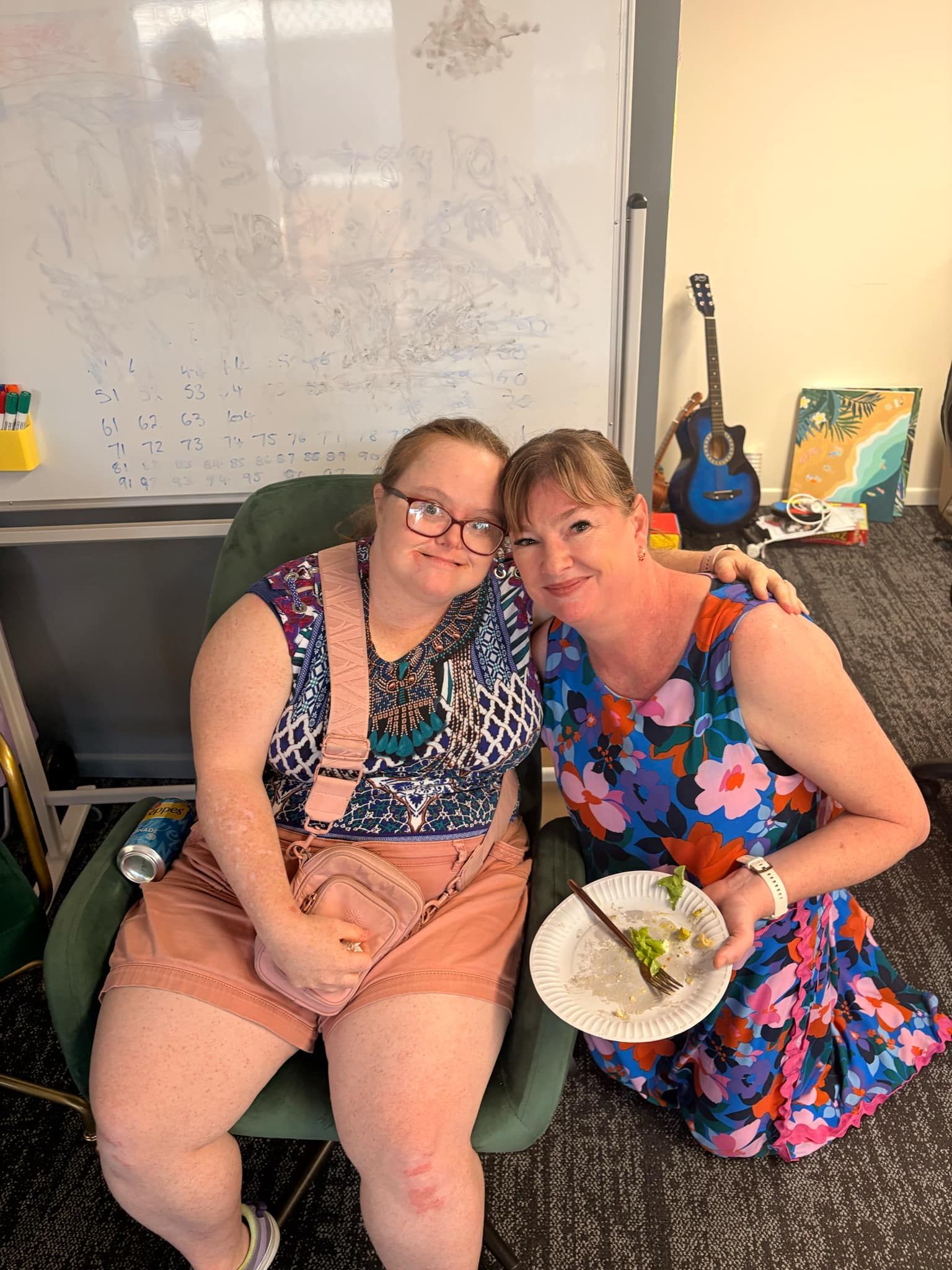 Two People Sit Together, One In A Green Chair And One Beside It Holding A Plate, Smiling In An Office With A Whiteboard — Advanced Therapy Mackay In Mackay, QLD 