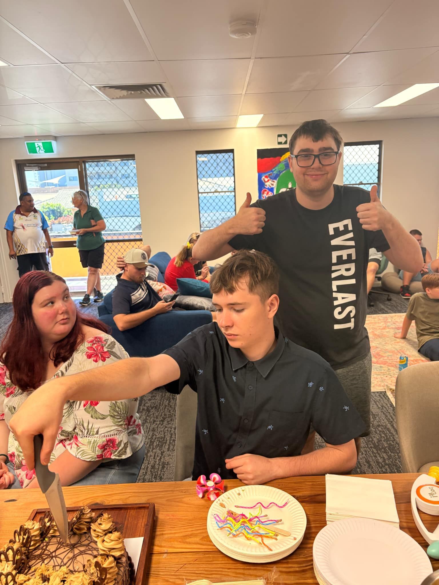 People gathered around a wooden table in a room, with one person cutting a cake while others look on or give a thumbs up — Advanced Therapy Mackay In Mackay, QLD 