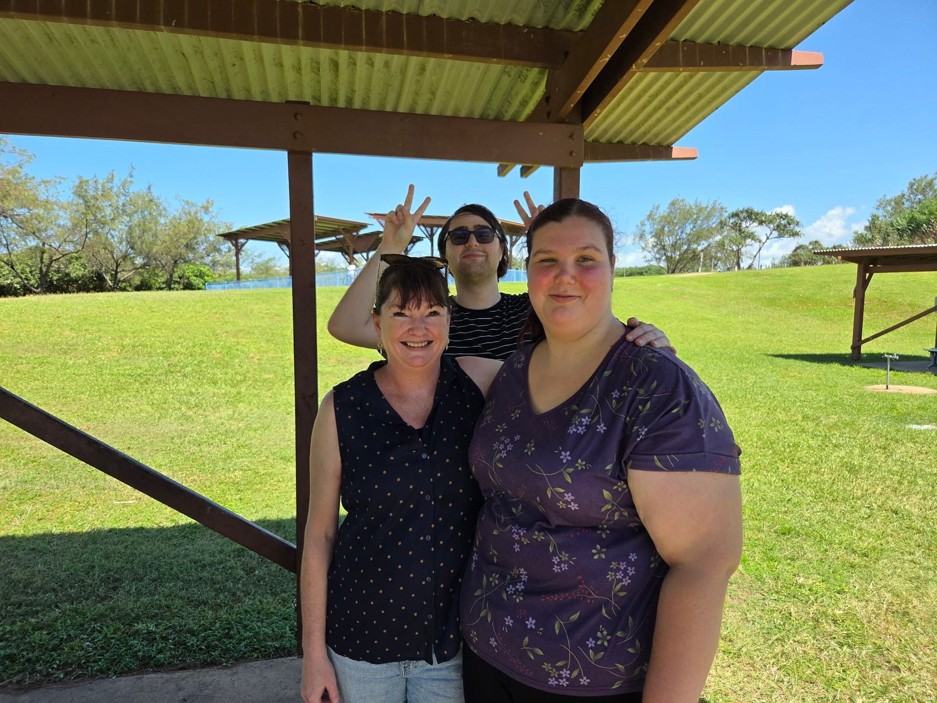 Three People Stand Smiling Under A Wooden Shelter On A Sunny, Grassy Day; One Person Holds Up Peace Signs Behind The Others — Advanced Therapy Mackay In Mackay, QLD 