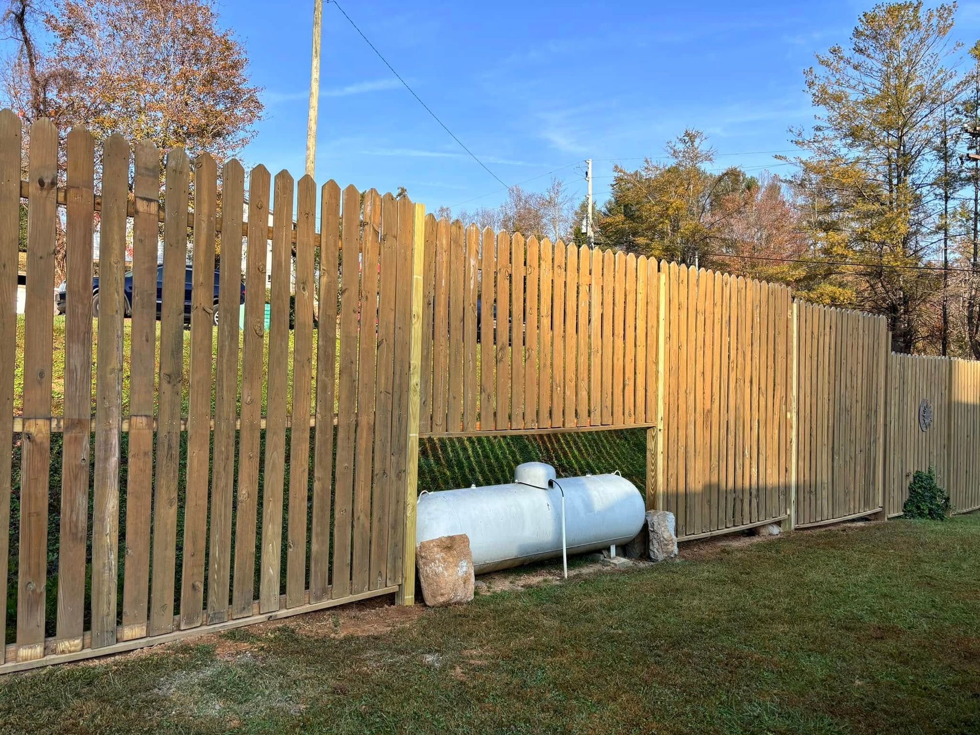 A large, white propane tank sits on a grassy lawn behind a gap in a wooden picket fence.