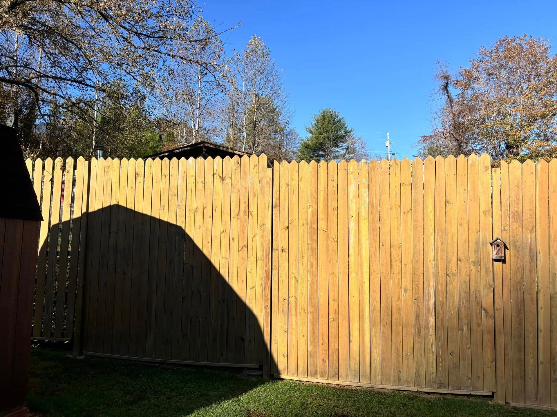 A wooden fence stretches across a backyard under a clear blue sky, with a dark shadow cast over part of the panels.