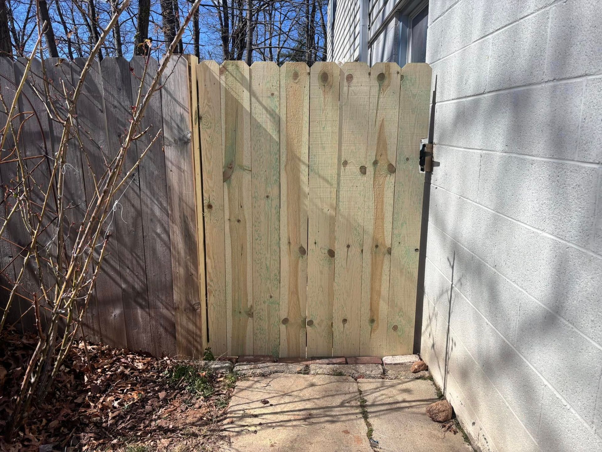 A new, light-colored wooden gate stands between a weathered wooden fence and a gray concrete block wall outdoors.
