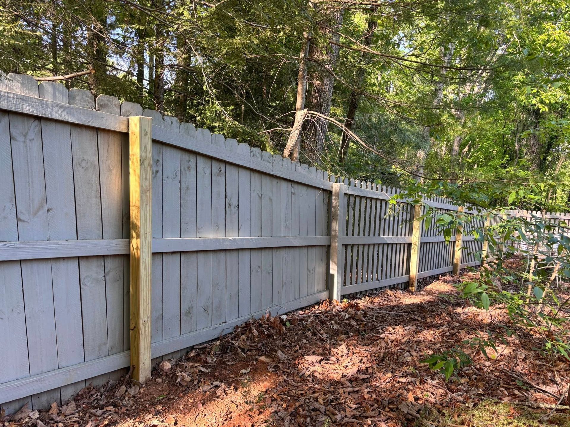 A gray wooden fence with new, light-brown vertical support posts stands in front of a forest with scattered fallen leaves.