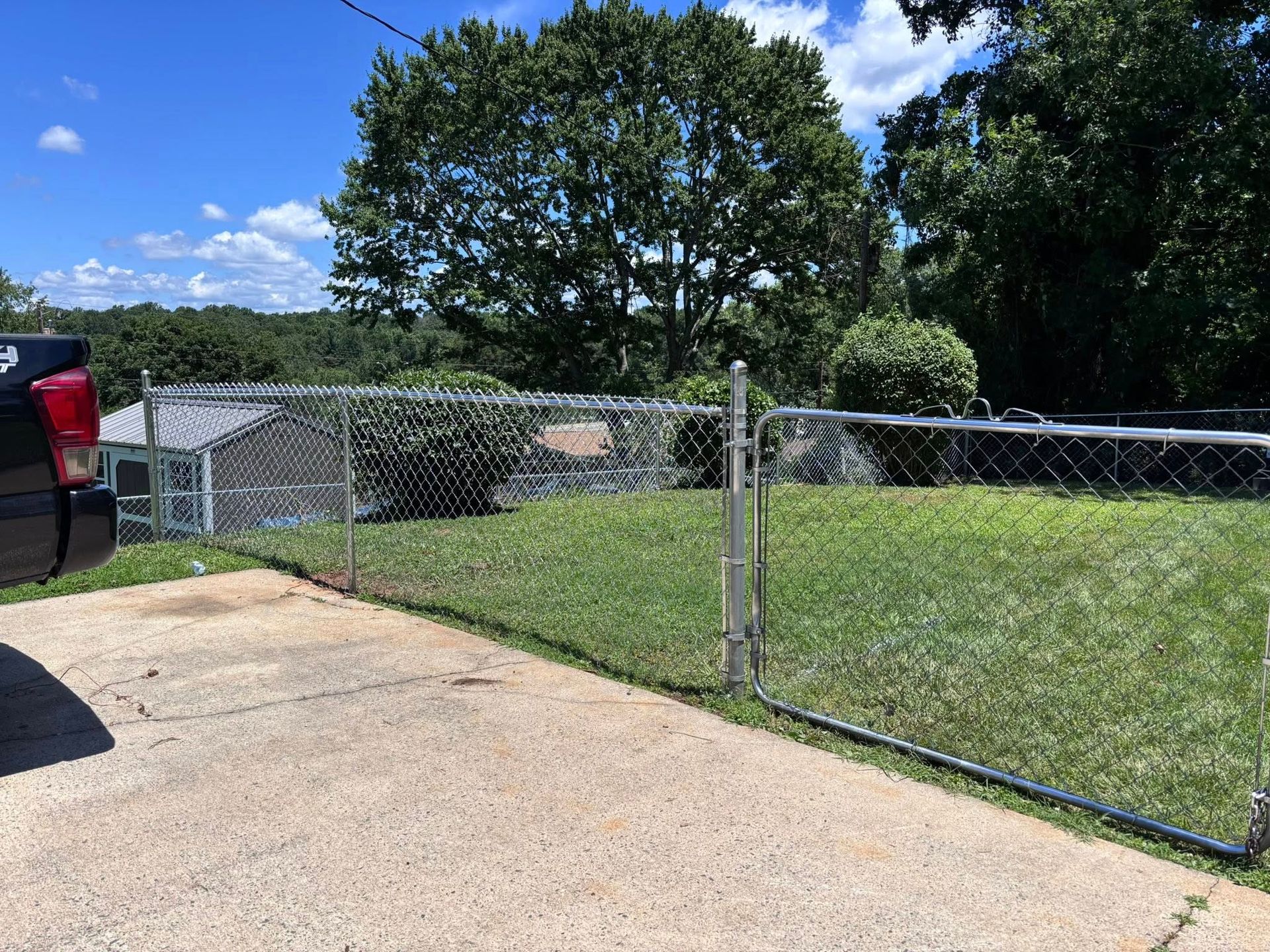 A driveway meets a chain-link fence and gate, opening onto a grassy yard with trees and a blue sky in the background.