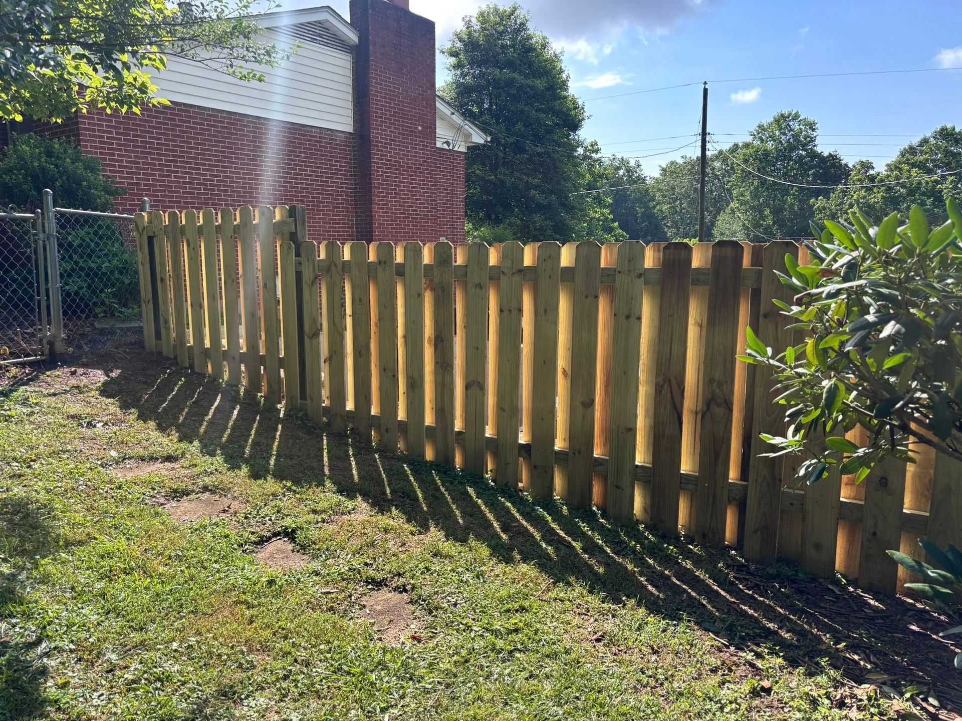 A new, light-colored wooden picket fence runs across a sunny, grassy backyard, positioned in front of a brick house.