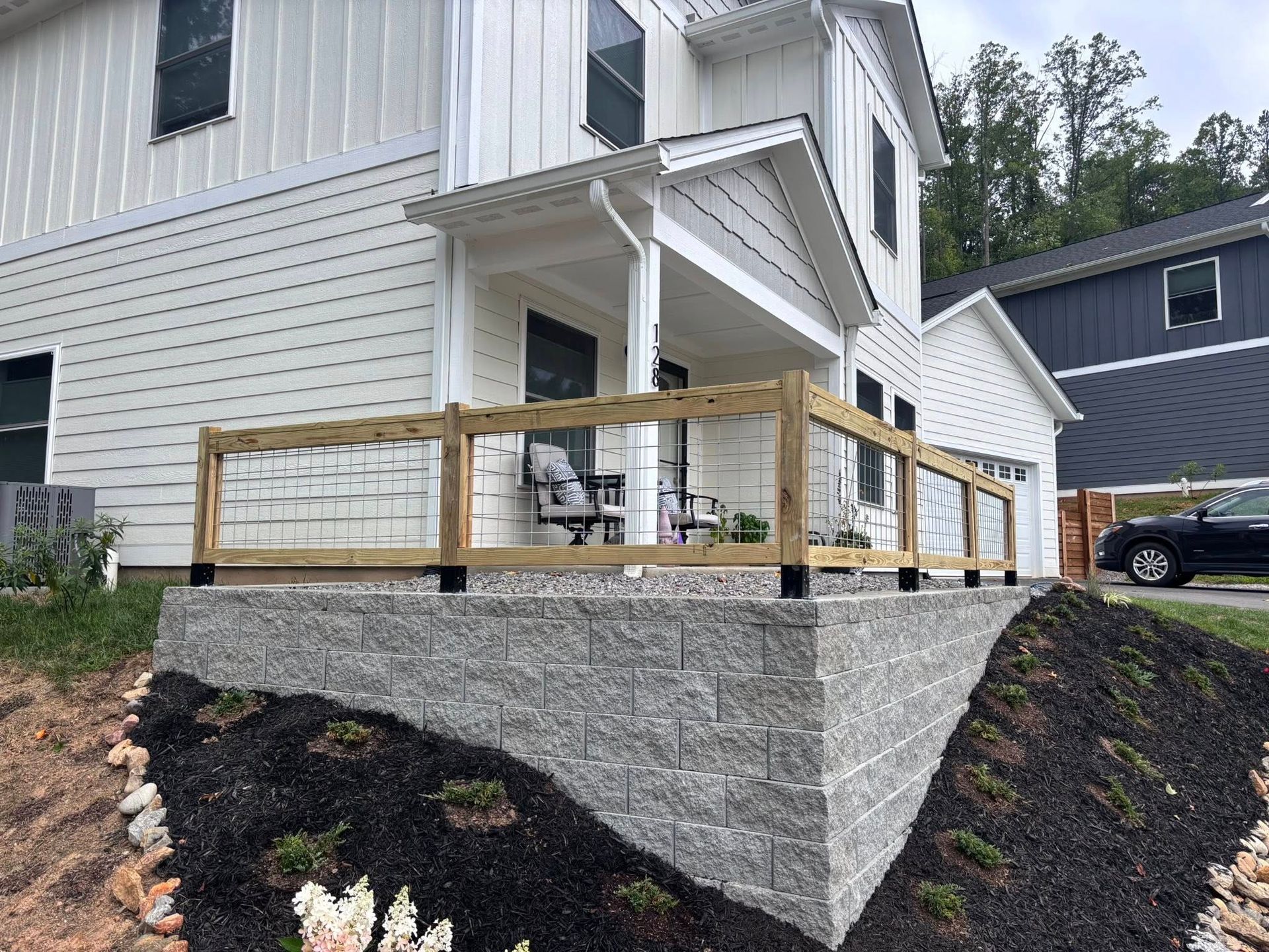 A modern white house with a raised porch featuring a stone retaining wall and a wooden railing with wire mesh infill.
