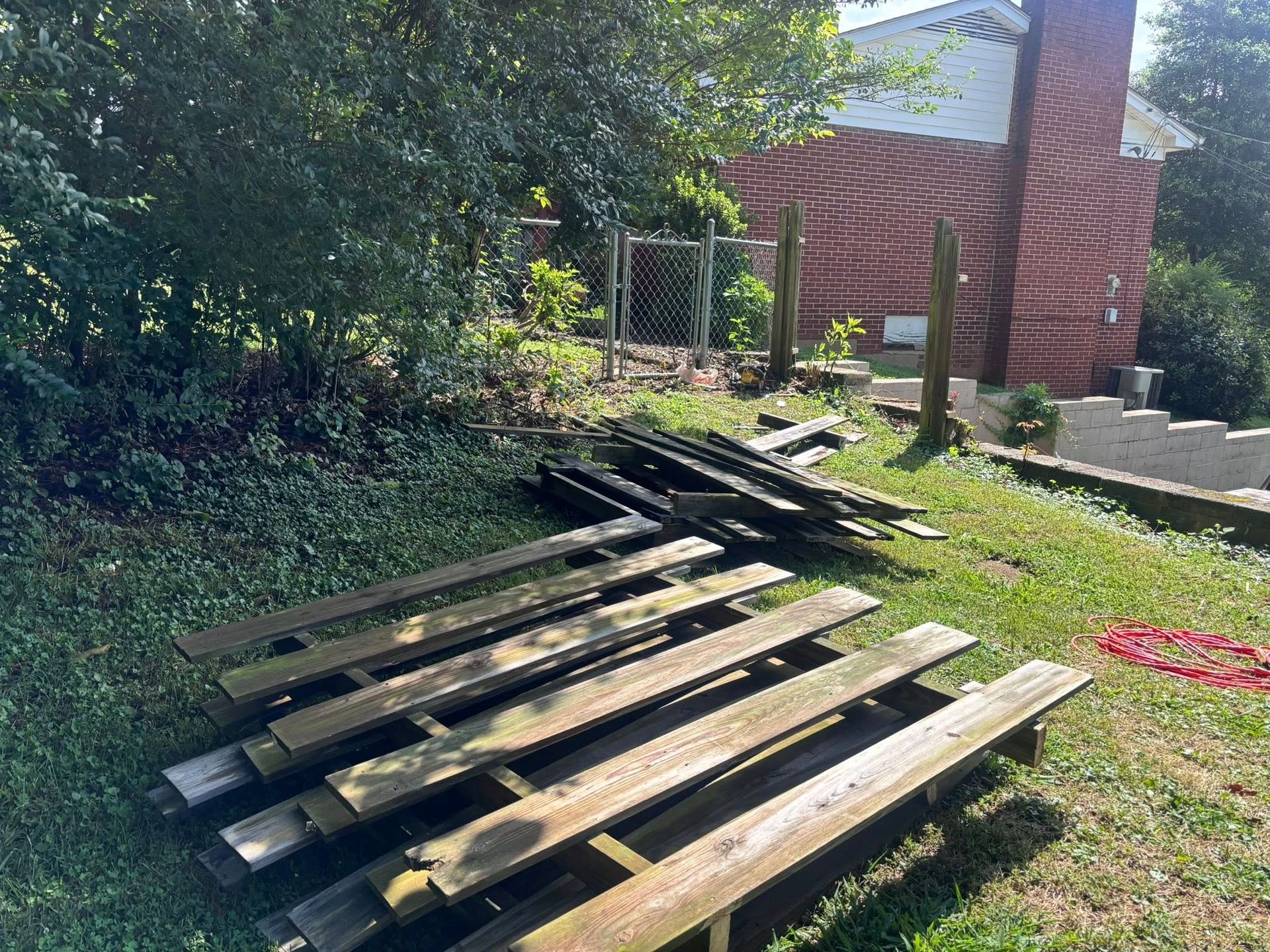 A pile of weathered wooden deck boards lies on a grassy lawn next to a brick house and a chain-link fence.