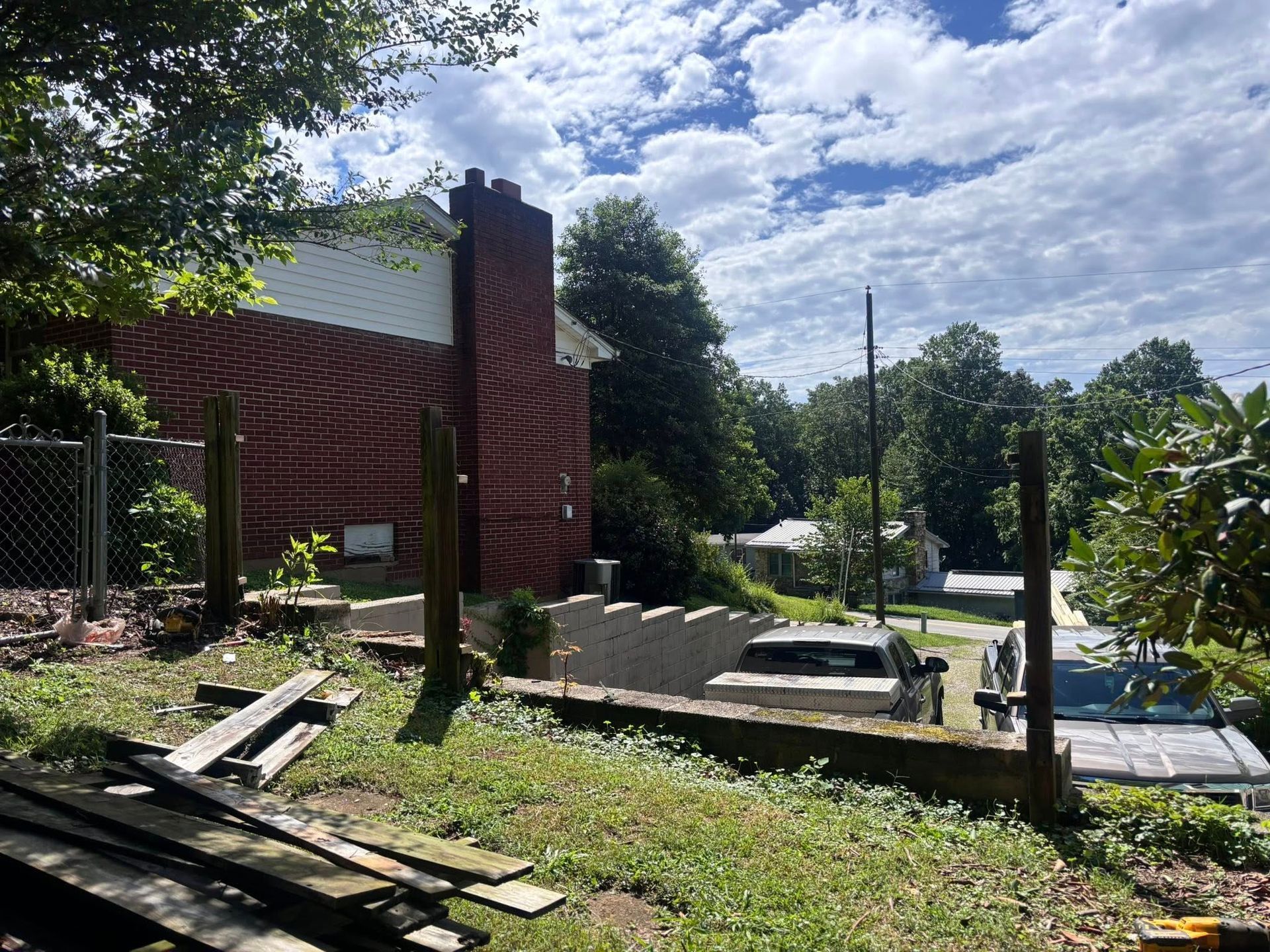 A brick house and a terraced backyard with wooden fence posts, stacked lumber, and parked cars under a cloudy sky.