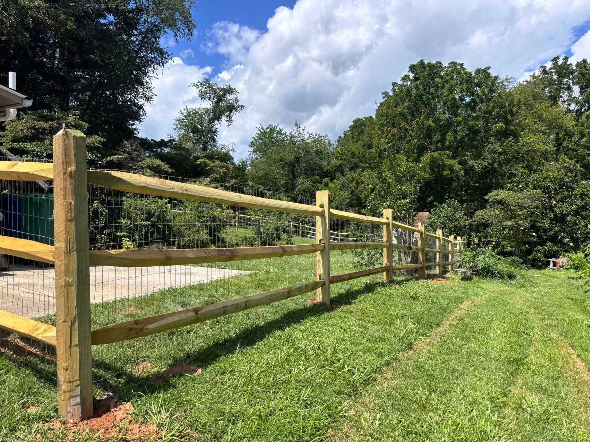 A wooden split-rail fence runs across a grassy lawn on a sunny day with trees in the background.