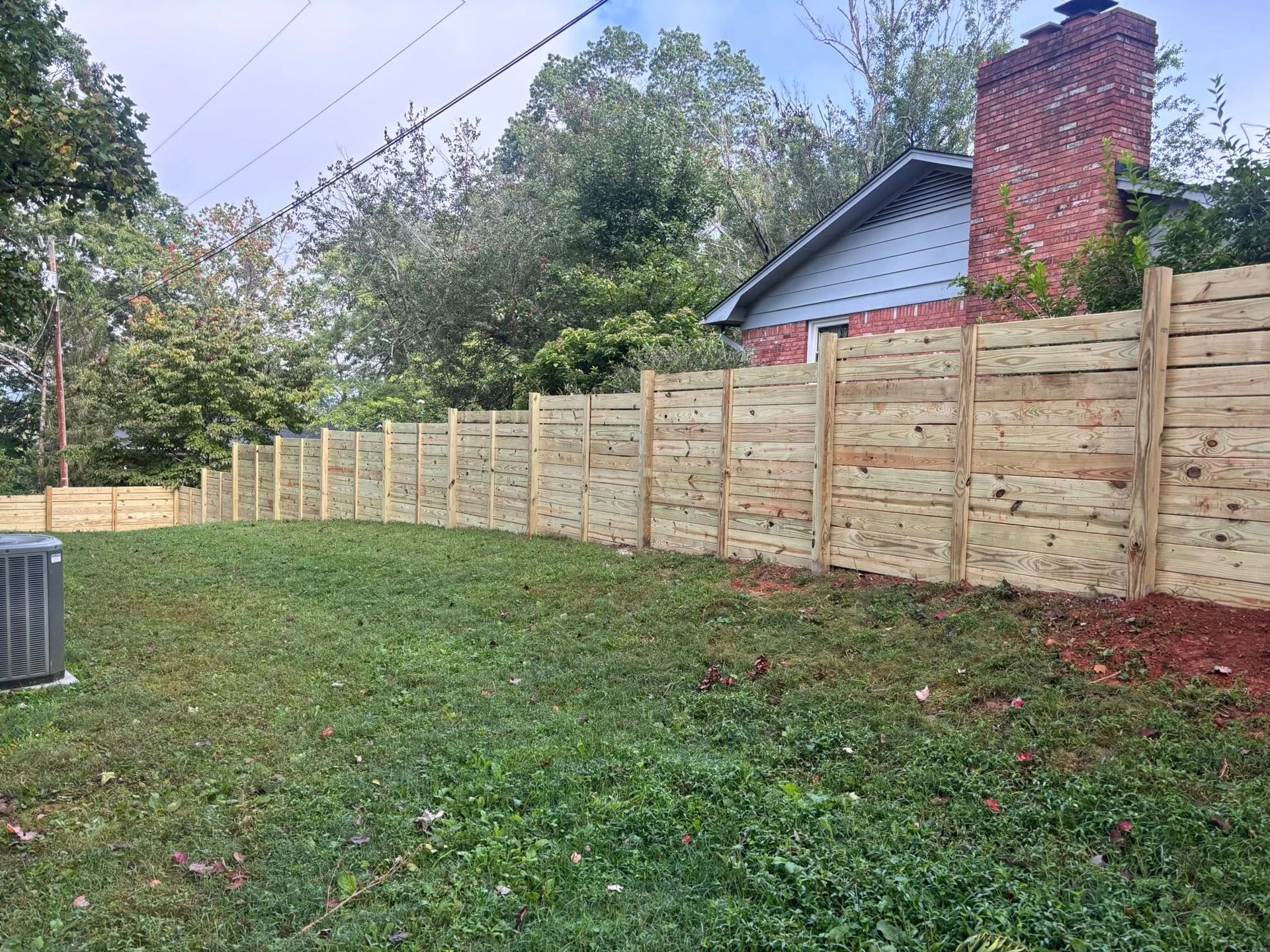 A new horizontal wooden privacy fence stretches across a grassy backyard, adjacent to a brick house with a blue gable.