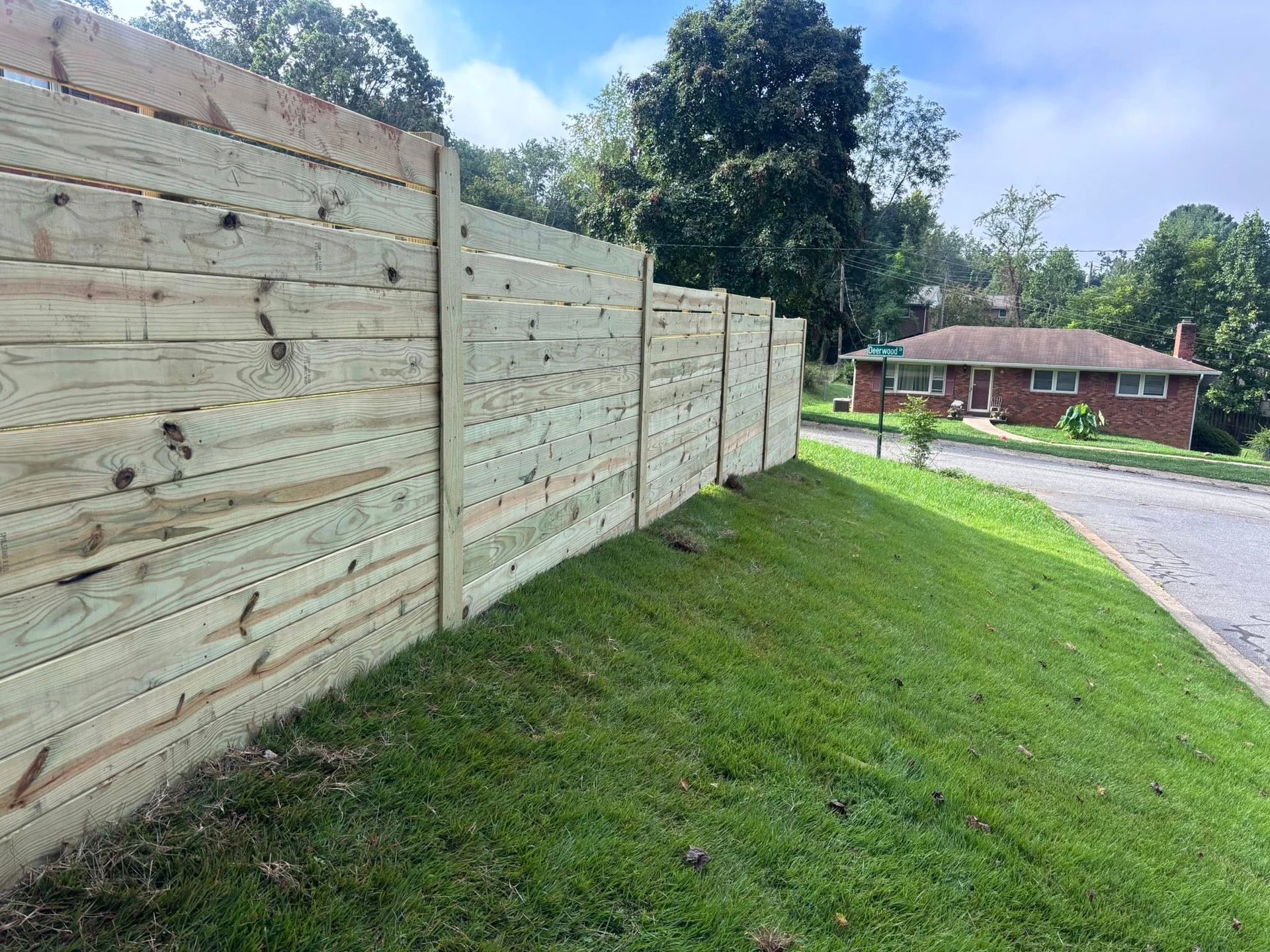 A tall, horizontal wooden fence runs along a grassy slope next to a road with a brick house in the background.