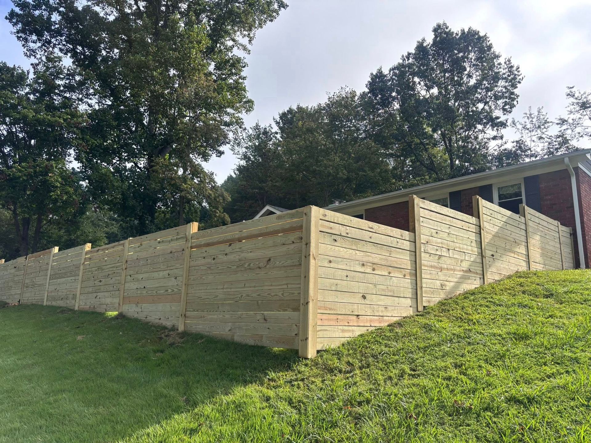 A light-colored wooden horizontal-slat fence stands on a grassy hill next to a brick house under a blue sky.